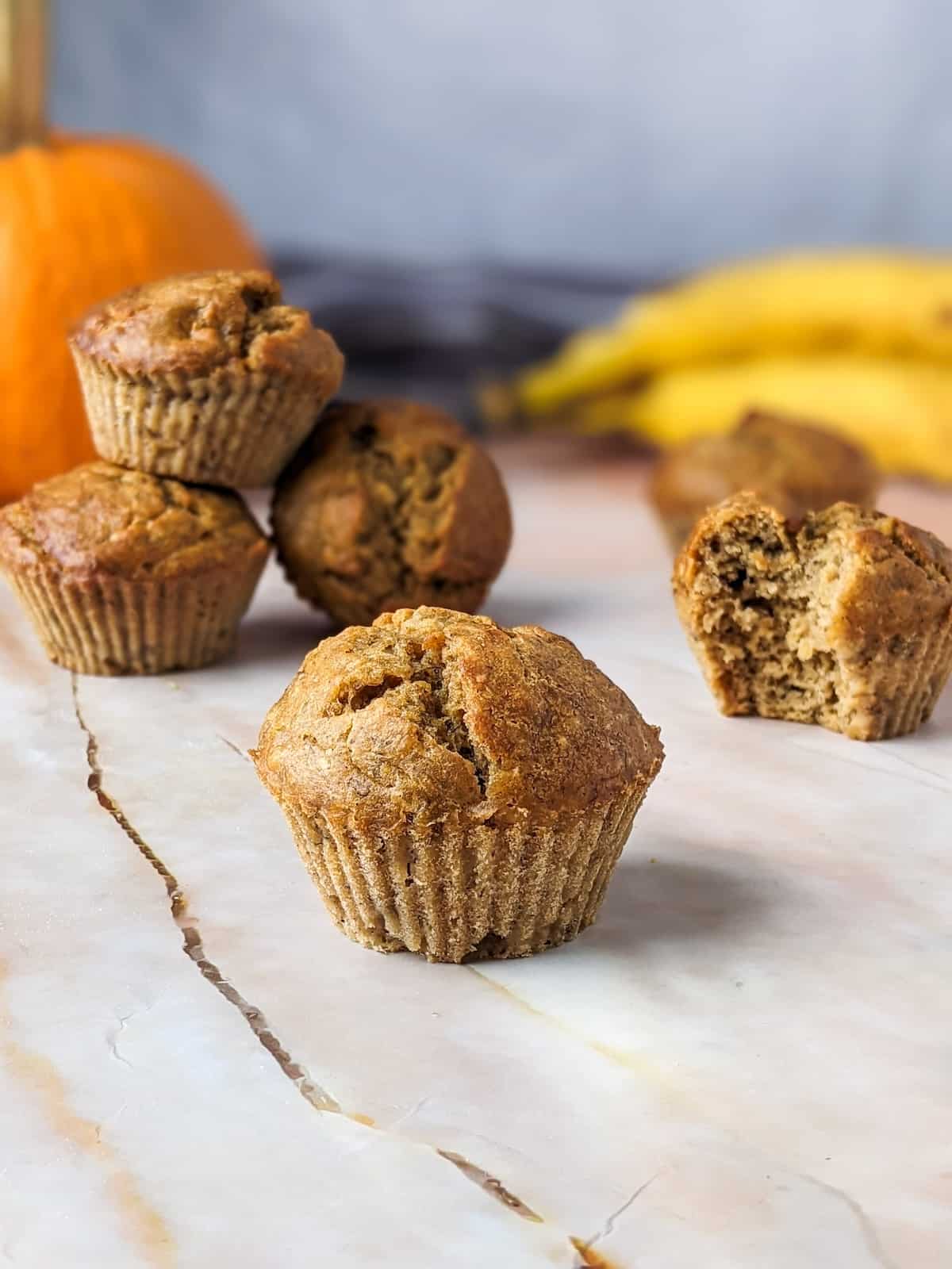 Pumpkin banana muffins on the table with a pumpkin and some bananas in the background.