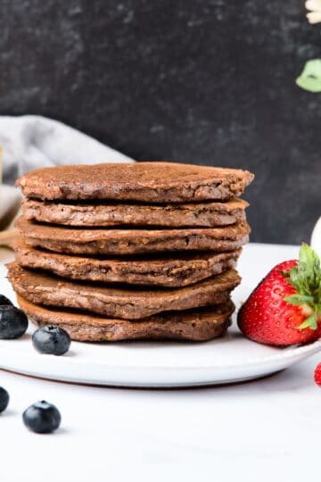 Chocolate oat pancakes on a white plate next to blueberries and strawberries.