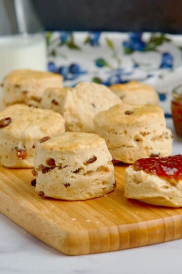 Fruit scones on a wooden board next to a jar with jam and a jug of milk.