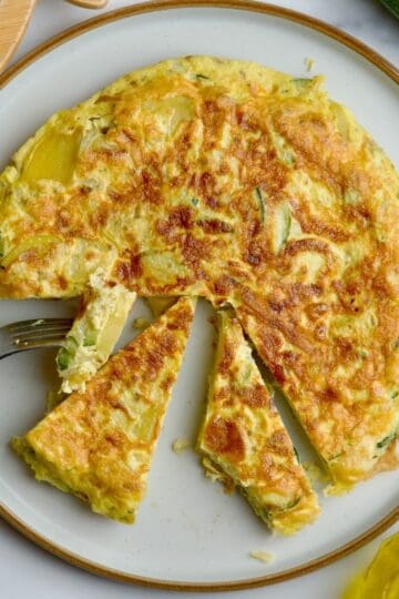 Potato and zucchini frittata served on a white plate next to olive oil and zucchini.