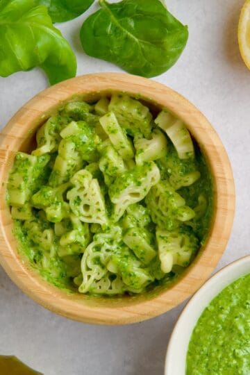 Pesto sauce with pasta in a bowl next to olive oil, lemons, green leaves and a bowl with pesto sauce.