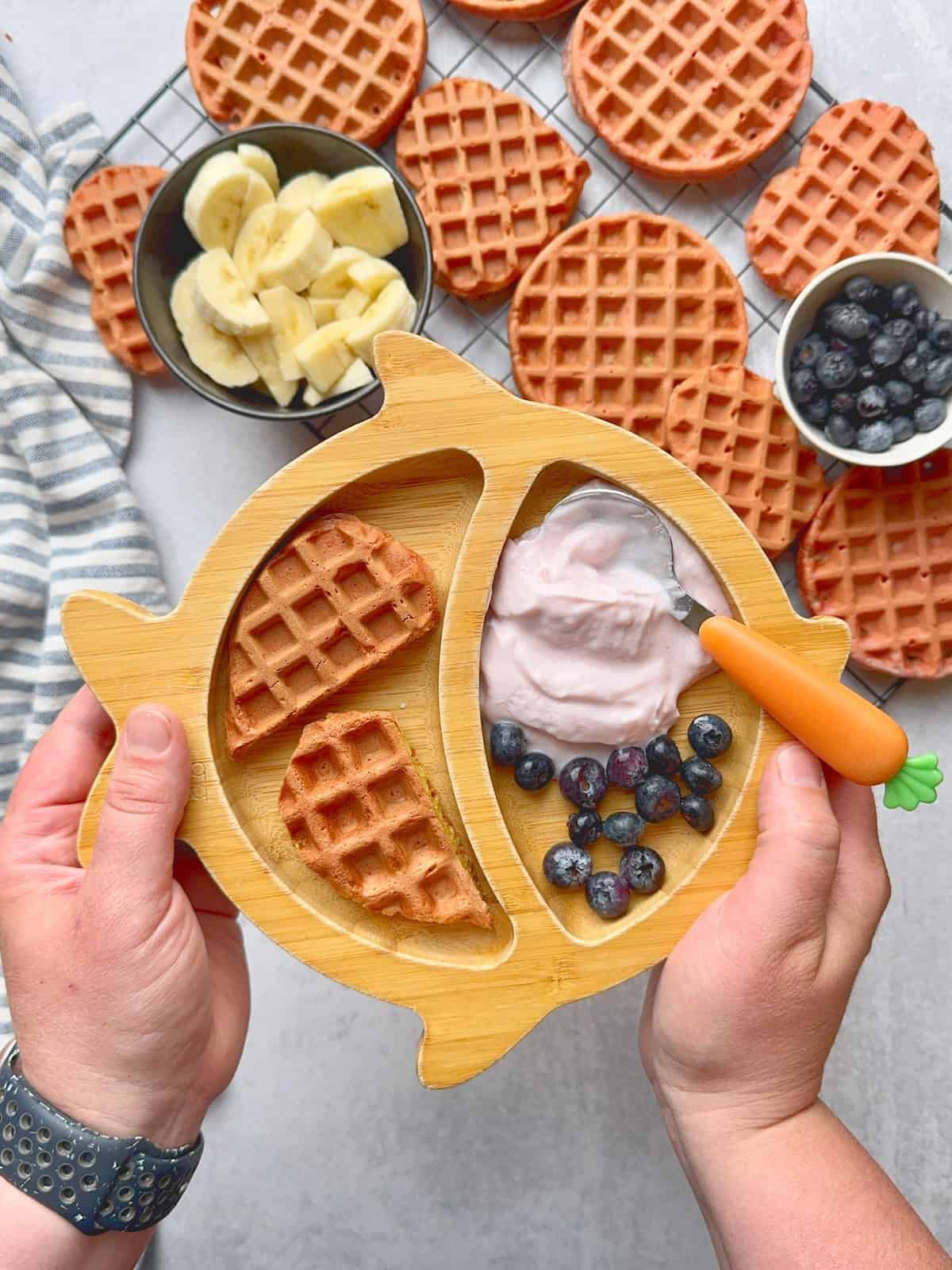 Hands holding bamboo plate with beet heart waffle, yogurt and blueberries.