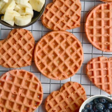 Beet waffles on a wire rack between a bowl of blueberries and bowl with banana slices.