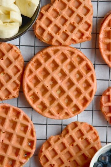 Beet waffles on a wire rack between a bowl of blueberries and bowl with banana slices.
