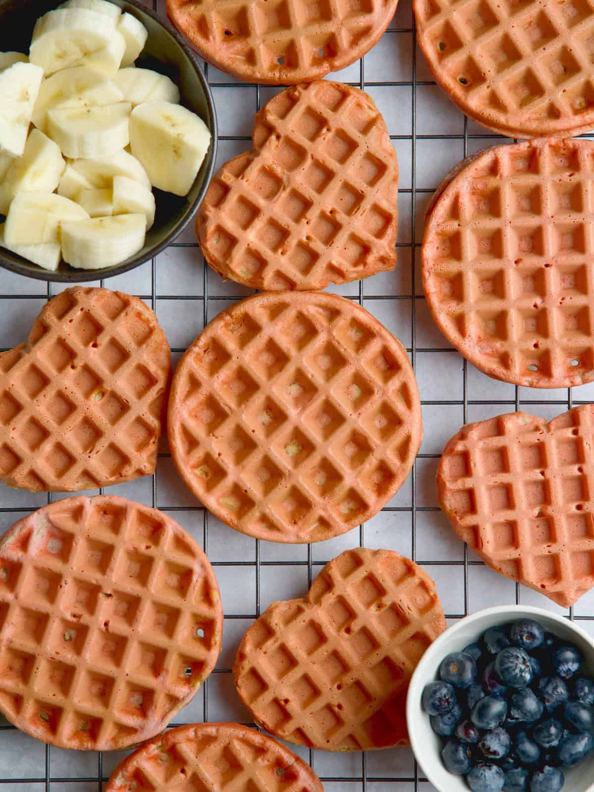 Beet waffles on a wire rack between a bowl of blueberries and bowl with banana slices.