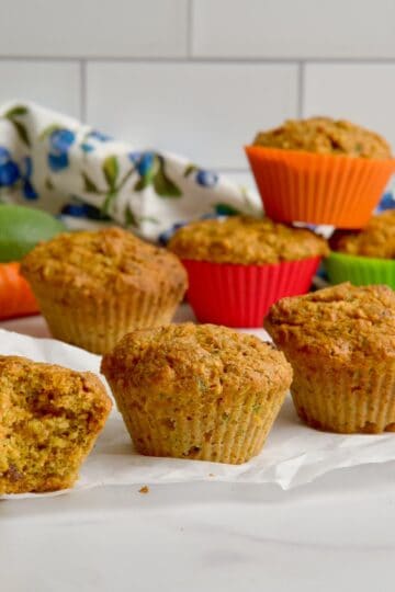 Morning glory muffins on the table with a zucchini and a carrot in the background.